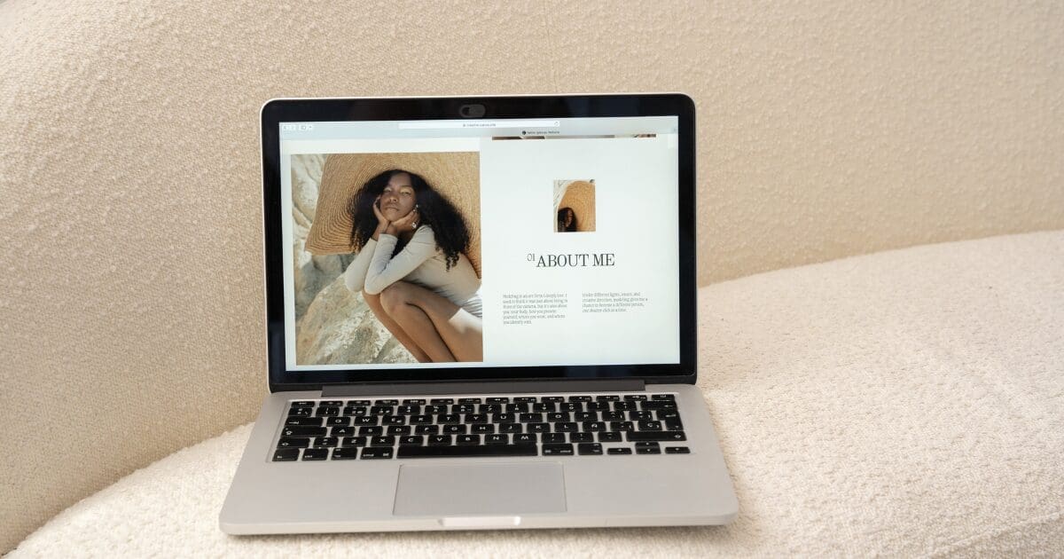 A MacBook laptop open on a neutral-toned surface displaying an "About Me" webpage layout, featuring a large photo of a woman in a wide-brim straw hat seated in a relaxed pose against a light background, alongside a smaller thumbnail image and editorial-style text columns.