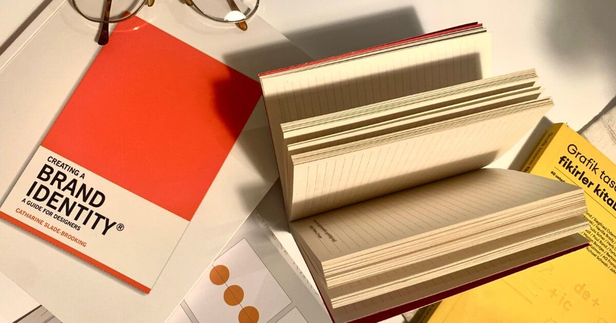 A flat lay of design books on a white surface, including "Creating a Brand Identity: A Guide for Designers" by Catharine Slade-Brooking with a bold orange cover, an open notebook with lined pages fanned upright, and a yellow book partially visible. A pair of round glasses rests in the upper left corner.